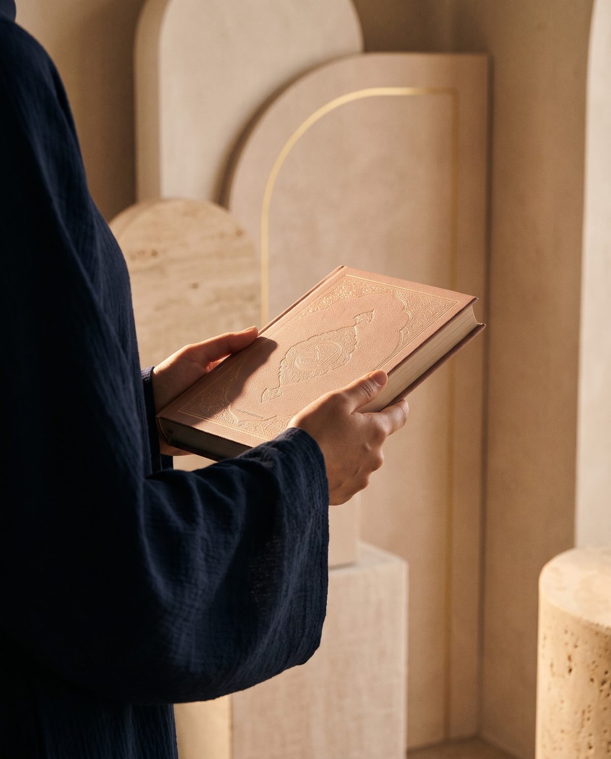 A woman in a navy abaya holding a closed rose-covered Quran, beside cream stone arches with a fine gold line